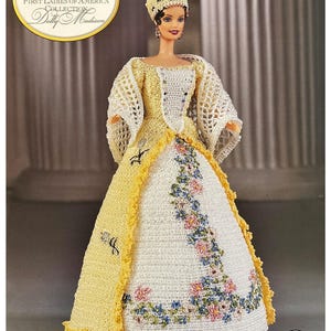 Könnte beinhalten: Eine Barbie-Puppe aus der First Ladies of America Collection, Dolly Madison. Die Puppe trägt ein gehäkeltes gelb-weißes Kleid mit floraler Stickerei und eine passende Haube. Die Puppe hat dunkles Haar und posiert vor einer Säule.