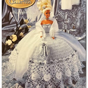 May include: A doll from the "Gems of the South Collection" named "Miss April" is featured. The doll is wearing a white crocheted gown with a matching shawl and a blonde updo. The image has a vintage aesthetic.