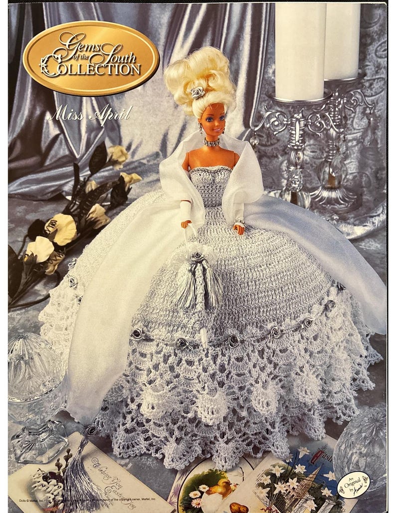 May include: A doll from the "Gems of the South Collection" named "Miss April" is featured. The doll is wearing a white crocheted gown with a matching shawl and a blonde updo. The image has a vintage aesthetic.