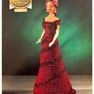 Könnte beinhalten: Ein rotes und schwarzes Häkelkleid mit einer weißen Spitzenborte. Das Kleid wird von einer Puppe mit braunen Haaren und einer Perlenkette getragen. Die Puppe steht auf einem grünen Hintergrund. Das Bild stammt aus der Golden Age of Film Collection.