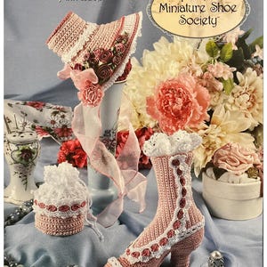 Op de afbeelding: Een verzameling miniatuur gehaakte schoenen en accessoires in roze en wit. De afbeelding toont een gehaakte laars, een hoed en een kleine tas, allemaal met delicate kant- en bloemendetails. De tekst luidt "Stepping Through Time Collection Pink Lace".