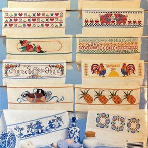 May include: A collection of embroidered decorative towels with various designs. Some towels feature floral patterns, while others display text like "Welcome" and "Home Sweet Home." The towels are displayed on a metal rack.