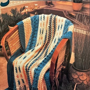 May include: A crocheted throw blanket in shades of blue, orange, and white with fringe on the edges. The blanket is draped over a wicker chair in a bathroom setting.