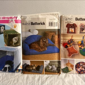 May include: Three sewing pattern envelopes featuring dog-related projects. One shows dog crates, another a dog on a blue sofa, and the third includes dog beds and Christmas stockings. The patterns are from Simplicity and Butterick.