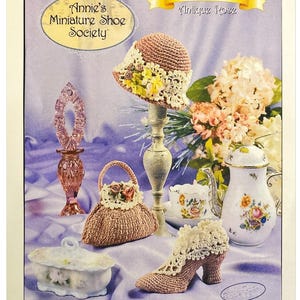 May include: A collection of miniature crochet items, including a hat, purse, and high-heeled shoe, arranged on a purple surface. The image features a decorative perfume bottle, a teacup, and a teapot. The text "Annie's Miniature Shoe Society" is visible.