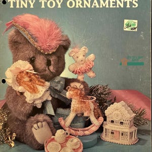 Puede incluir: Un libro de patrones de ganchillo vintage con 10 adorables adornos de juguete en miniatura. La portada muestra un oso de peluche, un caballito de balancín, una muñeca y una casa de ganchillo. El título del libro es "Victorian Crochet Tiny Toy Ornaments".