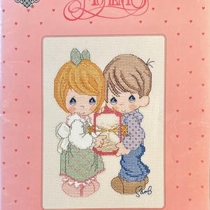 Puede incluir: Un kit de punto de cruz con dos figuras de Precious Moments sosteniendo una caja en forma de corazón. El kit incluye el texto "Counting Our Moments Together" y el logo de "Precious Moments". El fondo es rosa con pequeños corazones.