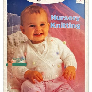 May include: A white knitting pattern book with a photo of a baby wearing a white knitted cardigan with colorful buttons. The book is titled "Nursery Knitting" and is by Susan Bates.
