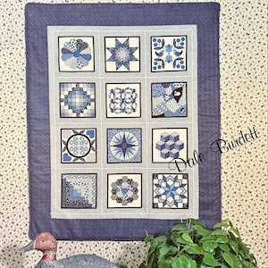 May include: A cross-stitch quilt sampler with a blue border and a floral background. The sampler features twelve square designs in shades of blue, with the text "the Cross Stitch Quilt Sampler" at the top. The text "3 Color Schemes Offered" is also visible.