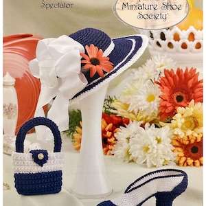 May include: A navy blue and white crocheted hat with a white bow and orange flower sits on a white stand. A matching purse and high-heeled shoe are also displayed. The image includes text that reads "Annie's Miniature Shoe Society."