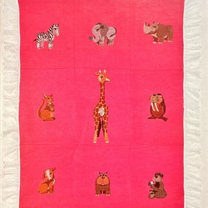 May include: A bright pink quilt featuring a grid of nine embroidered animal figures including a zebra, elephant, rhinoceros, kangaroo, giraffe, walrus, goat, bulldog, and bear. The quilt has a white ruffled border and text reading Mary Ellen Designs Wild and Woolies Friendly Creatures.