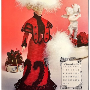 May include: A crocheted doll wearing a red dress with black trim and a white feathered hat. The doll is holding a white feathered fan. The doll is standing in front of a calendar for December 1996.