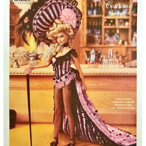 Puede incluir: Una muñeca con un disfraz de ganchillo del patrón "Crochet Collector Costume Volume 87". La muñeca lleva un bustier negro y rosa, una larga cola y un gran sombrero con plumas y una flor rosa. La imagen incluye el texto "1899 WILD WEST SHOWGIRL".