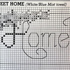 May include: Cross-stitch pattern with the words "HOME SWEET HOME" in a block letter font. The design includes decorative elements like stars and geometric shapes. The text "(White/Blue Mist towel)" is above the main text.