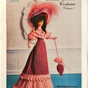 May include: A doll wearing a crochet costume, including a wide-brimmed hat with pink feathers, a long dress, and a parasol. The image is from a "Crochet Collector Costume" pattern, volume 5, and includes the text "1903 Promenade Dress."
