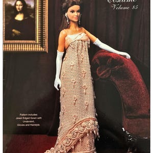 May include: A doll wearing a crocheted gown with a train, long white gloves, and a satin top. The cover of a "Crochet Collector Costume" book is visible, with the text "1963 Jeweled Presidential Gown." The doll is posed near a red velvet chair.