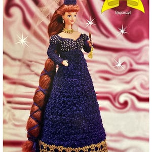 Pode incluir: Uma boneca de crochê vestida como Rapunzel da coleção de contos de fadas. A boneca está usando um vestido roxo com detalhes dourados, um colar de ouro e um chapéu de crochê. Seu cabelo comprido está trançado e amarrado com uma fita. A boneca está segurando um espelho em sua mão direita.