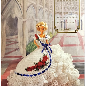 Puede incluir: Un vestido de muñeca de crochet blanco con rosas rojas y detalles de cinta azul. El vestido lo lleva una muñeca rubia con una banda azul. El fondo es una escena pintada de un edificio con arcos. El calendario de julio de 1992 es visible en la esquina superior derecha.
