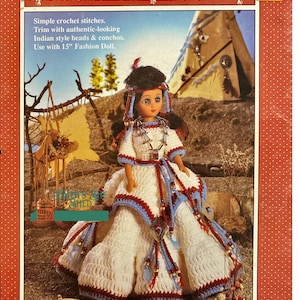 Könnte beinhalten: Ein Häkelmuster für ein Indianer-Prinzessinnen-Puppenkostüm. Die Puppe trägt ein weißes Kleid mit roten und blauen Verzierungen, Perlenketten und einen Federkopfschmuck. Das Muster ist von Fibre Craft.