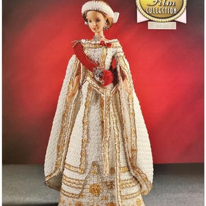 May include: A white and gold doll wearing a crown and a red sash. The doll is part of the "The Golden Age of Film Collection" and is dressed as a queen.
