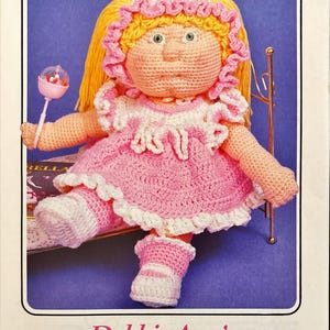 May include: A crocheted doll wearing a pink dress with white lace trim and white booties. The doll has blonde hair and blue eyes. The doll is sitting on a bed with a gold metal headboard. The doll is holding a rattle in its hand. The image is from a crochet pattern book.