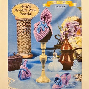 Puede incluir: Exhibición de artículos de ganchillo en miniatura en rosa y azul, incluyendo un sombrero, un bolso y un zapato. Los artículos están dispuestos sobre una superficie azul con elementos decorativos. El texto "Annie's Miniature Shoe Society" es visible.