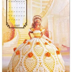 May include: A crochet doll wearing a white and yellow dress with orange flowers. The doll is standing on a yellow and white background. The image includes a calendar for April 1992.