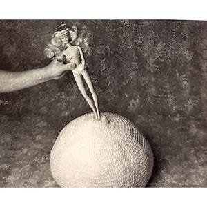 May include: Black and white image of a blonde doll standing on a textured, spherical object. The doll is held by a hand, and the background is a mottled gray. The doll is wearing a white outfit.