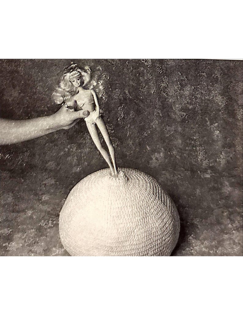 May include: Black and white image of a blonde doll standing on a textured, spherical object. The doll is held by a hand, and the background is a mottled gray. The doll is wearing a white outfit.