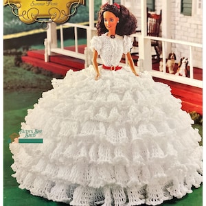 May include: A white crochet doll dress with multiple layers of ruffles. The dress is worn by a Barbie doll with brown hair and a red bow. The doll is standing on a porch with a rocking chair and a small dog.