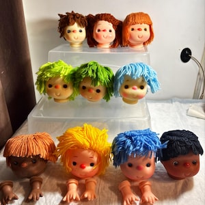 May include: A collection of eleven doll heads with yarn hair in various colors including brown, orange, green, blue, yellow, and black. Each head features a painted face with rosy cheeks, freckles, and a small nose. Some heads include matching plastic hands.