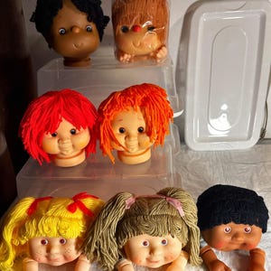 May include: Seven doll heads with yarn hair in various colors including black, orange, red, yellow, and brown. The dolls feature painted facial details, including eyes, noses, and mouths, with some having freckles. One doll head is inside a plastic bag.