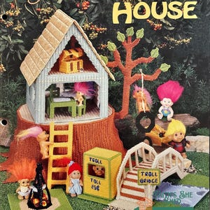 May include: Plastic canvas needlepoint pattern for a troll house with a tree, a bridge, a toll booth, and several trolls. The pattern is titled "Troll House" and is by Karen McDanel.