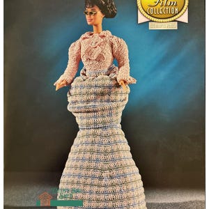 May include: A crochet doll wearing a pink and blue dress with a matching hat. The doll is part of the "The Golden Age of Film Collection" and is titled "The Movie Star"