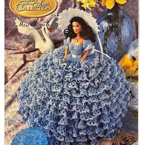 May include: A crochet doll pattern for "Miss March" from the "Gems of the South Collection". The doll is wearing a blue and white crocheted dress and a white crocheted hat. The doll is sitting on a floral patterned fabric.