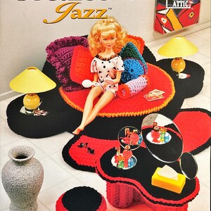 May include: A doll sitting on a crocheted red and black rug with pillows and a table. The rug and table are decorated with a black and red color scheme. The image also includes a vase, a lamp, and the text "Bedroom Jazz".