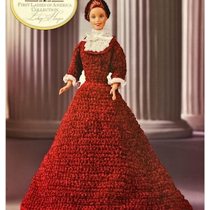 Può includere: Bambola della collezione First Ladies of America, Lucy Hayes, indossa un abito rosso all'uncinetto con finiture in pizzo bianco. La bambola ha i capelli rossicci e posa davanti a uno sfondo sfocato con colonne. La bambola è un originale di Mattel.