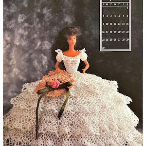 May include: A white crocheted doll dress with a ruffled skirt and a matching crocheted hat. The doll is wearing a white crocheted bodice with a lace trim. The doll has brown hair and blue eyes.