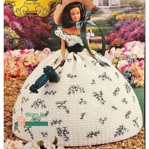 Op de afbeelding: Een gehaakte pop die een witte jurk draagt met groene bloemenborduursels, een groene hoed en een groene parasol. De pop staat in een tuin met bloemen en een prieel op de achtergrond. De pop maakt deel uit van de "Southern Belle Collection" en heeft de titel "Lawn Party".