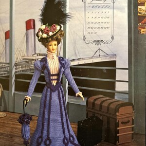 May include: Crocheted doll wearing a purple dress with a pink and white blouse, a hat with a black feather, and holding a crocheted umbrella. The doll is standing in front of a vintage suitcase and a ship.