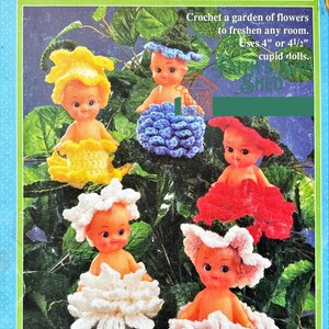 Op de afbeelding: Vijf gehaakte bloemenpoppen op een lichtblauwe achtergrond. De poppen zijn gekleed in gehaakte bloemen in tinten geel, blauw, rood, wit en roze. De poppen zijn 10 of 11,5 cm hoog. De tekst op de afbeelding luidt "Bud Babies Crochet a garden of flowers to freshen any room. Uses 4" or 4 1/2" cupid dolls."