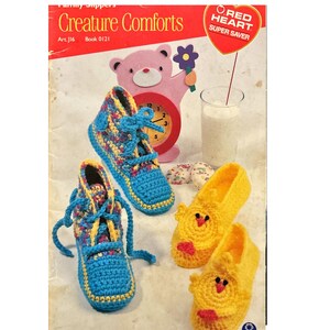 May include: A crochet pattern book for making creature-themed slippers. The cover features a pink teddy bear, a clock, a glass of milk, and two pairs of slippers. One pair is blue and yellow with laces, and the other pair is yellow with a chick design. The book is titled "Creature Comforts" and is a Red Heart Super Saver pattern.