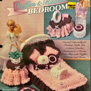 May include: A crochet pattern for a doll's bedroom set, featuring a bed, rug, and accessories. The set is in shades of pink, brown, and white, with ruffles and ribbons. The text on the pattern reads "Ruffles & Ribbons BEDROOM".