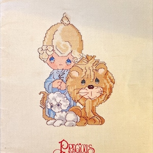 May include: Cross-stitch pattern featuring a child with blonde hair, a lion, and a small dog. The child wears a blue outfit and holds a sprig. The words "Peace on Earth" and "Precious Moments" are visible.