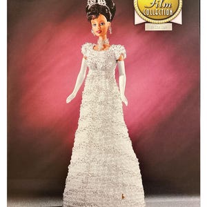 May include: A white, beaded, floor-length gown with a white lace bodice. The doll is wearing white gloves and a tiara. The doll has brown hair and brown eyes. The background is a pink gradient. The image is part of the "The Golden Age of Film Collection" series.