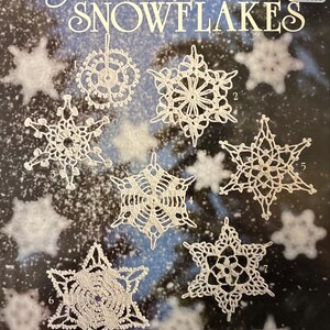May include: A craft instruction book titled Crocheted SNOWFLAKES by Mary Thomas, Book 1025(25). The cover features seven distinct white crocheted snowflake designs numbered one through seven against a dark blue background with white snowflake patterns.