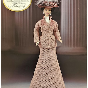 Puede incluir: Una muñeca Barbie de la colección First Ladies of America, Helen Taft, con un atuendo de ganchillo. La muñeca viste una falda larga marrón claro y una chaqueta a juego con un sombrero decorativo. El sombrero está adornado con plumas blancas.