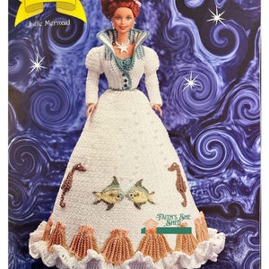 Puede incluir: Una muñeca de crochet vestida de sirena. La muñeca tiene el pelo rojo y lleva un vestido blanco con un corpiño verde. El vestido está decorado con caballitos de mar, peces y coral. La muñeca está de pie sobre un fondo azul con estrellas blancas. El texto "The Fairy Tale Collection" y "Little Mermaid" está en la parte superior de la imagen. El texto "Faith's She Shed" está en la parte inferior de la imagen.