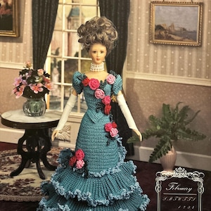 May include: A doll wearing a teal crocheted gown with pink and black floral accents. The gown has multiple layers of ruffles. The doll has brown hair styled in an updo and is wearing a pearl necklace.