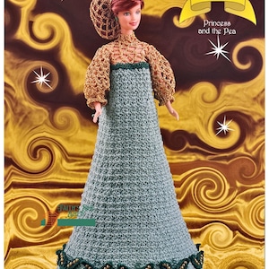 Puede incluir: Una muñeca de crochet vestida con un vestido verde y dorado con un sombrero de crochet. La muñeca forma parte de una colección llamada "The Fairy Tale Collection: Princess and the Pea".